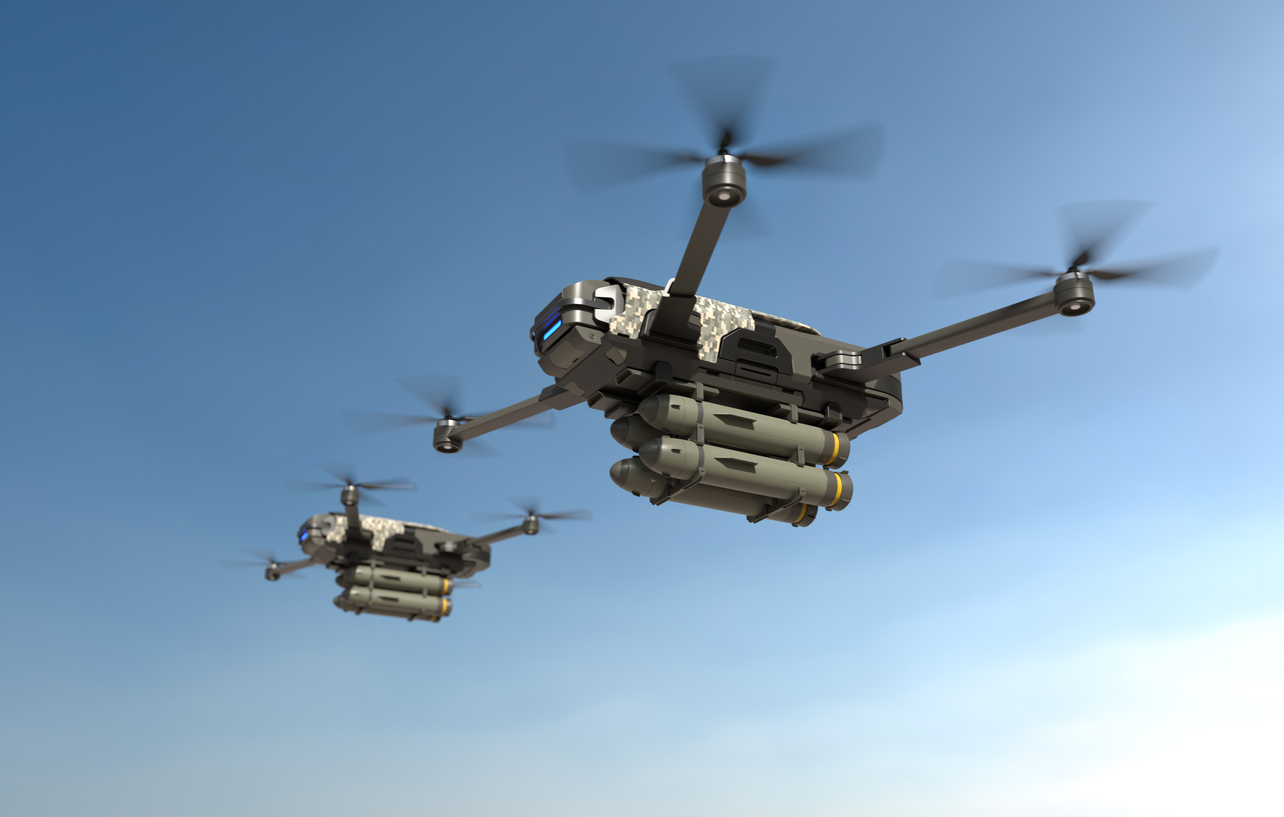 Two military drones carrying ordnance fly in a blue sky.